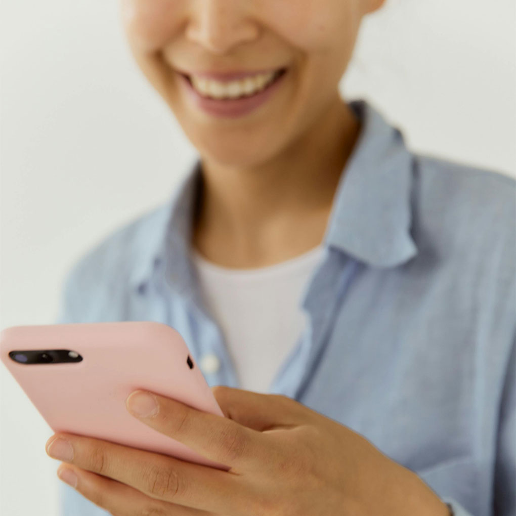 Close-up of a woman smiling while holding a pink smartphone in her hands.